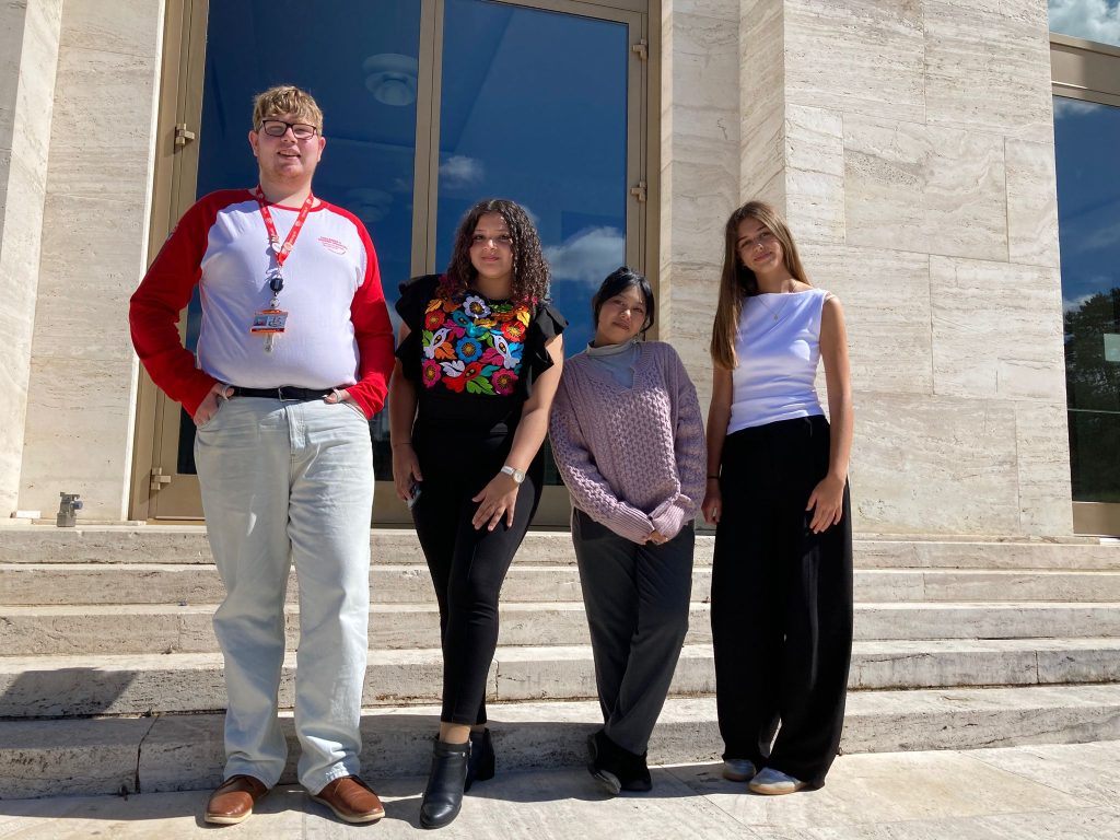Four young people stand together on stone steps in front of a large glass doorway. They are smiling slightly and dressed in casual clothes, with the sun casting strong shadows.