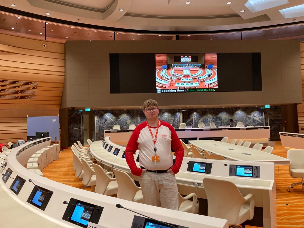 A young person stands inside a large United Nations conference room with curved rows of desks and screens. A big display screen shows a live view of the chamber, and the atmosphere is formal and official.