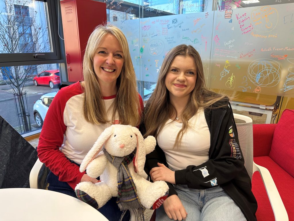 The Children and Young People’s Commissioner sits beside Young Advisor Niamh in a bright room, both smiling. The Commissioner is holding a soft white toy rabbit wearing a scarf. Behind them is a glass wall covered in colourful drawings and messages from children, with windows looking out onto parked cars in the background.